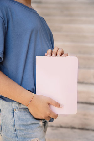 Kid girl in blue t-shirt standing and holding light pink notebook in hands close up, cropped shot. Notebook mockupの写真素材