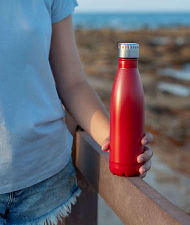 Young woman in light blue t-shirt holding red reusable water bottle in hand close up, cropped shot. Bottle and t-shirt mockupの写真素材