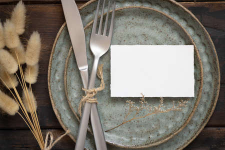 Blank white paper card on plate with fork and knife on wooden table with bohemian decoration and dried plants, top view. Boho wedding place card mockupの写真素材