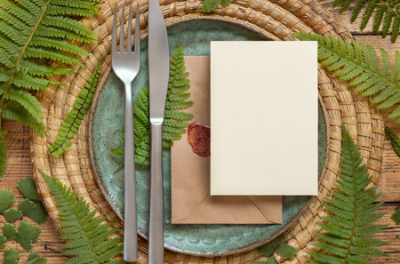 Blank paper card and sealed envelope on table setting decorated with fern leaves on wooden table top...