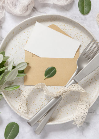 Blank paper card in envelope laying on white plate with fork and knife on marble table with eucalyptus branches and vintage ribbons around, top view. Card mockupの写真素材