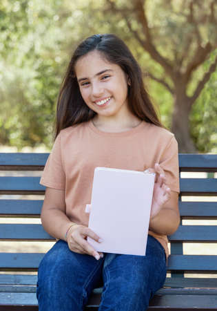 Kid girl in beige t-shirt sitting on a beench, holding notebook in hands and smiling, three quarter length. Notebook mockupの写真素材