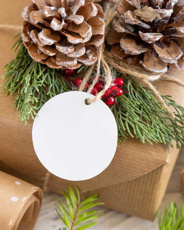 Christmas wrapped present with round paper gift tag on a white table with fir tree branches and decorations close up. Rustic winter composition with blank Gift tag Mockup, copy spaceの写真素材