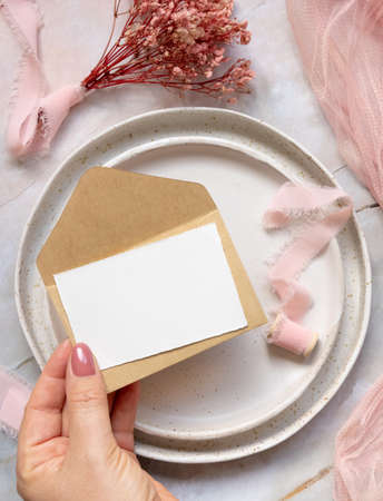 Hand with a card and envelope over wedding table place on a marble near dried pink flowers and silk ribbons top view. Flat lay with horizontal blank card. Romantic Thank you or rsvp card mockup, copy spaceの写真素材