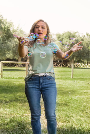 Women wearing t-shirt and jeans playing with soap bubbles in a park, three quarter shotMiddle aged...