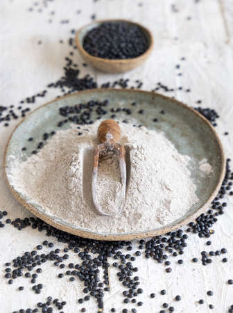 Plate with a scoop of black lentils flour and beans on white wooden table close up.  Alternative flour, healthy eating and vegetarian concept. Traditional Asian and North African cuisines ingredientの写真素材