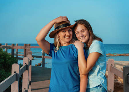 Mother wearing straw hat and teenage daughter standing on pier at sea coast, embracing and smiling, t-shirts mockup. Loving family spending time together outdoors in summer, enjoying sunset. Travel conceptの写真素材