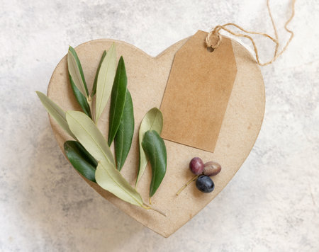 Kraft paper gift tag on a heart gift box on a white marble table with olive branches top view. Elegant modern template with vertical label tag. Mediterranean flat lay mockup with place for textの写真素材