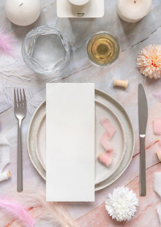 Wedding Table place with a paper card on porcelain plates near pink flowers, feathers  and silk ribbons top view menu mockup. Romantic template with vertical blank card flat lay. Invitation or menu card mockup, copy spaceの写真素材