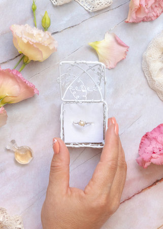 Woman hand opens a gift box with a pearl ring with light pink flowers and vintage ribbons around top...