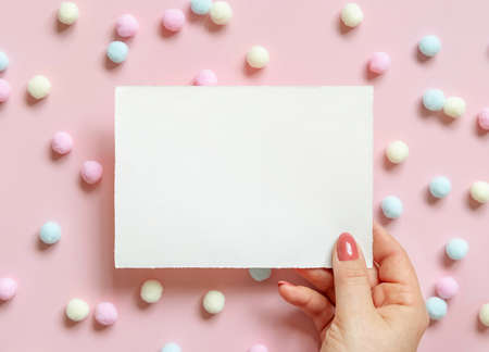 Hand with blank card near pastel pom-poms on pink top view. Romantic scene with blank horizontal card mockup flat lay, place for text . Valentines, Spring or girlish conceptの写真素材