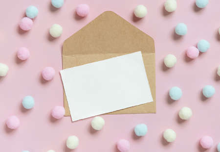 Blank card on envelope near pastel pom-poms on light pink top view. Romantic scene with blank horizontal card with place for text, flat lay. Valentines, Spring or girlish conceptの写真素材