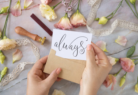 Hands with Card ALWAYS inside envelope near by pink flowers, wax seal and ribbons on a marble table...