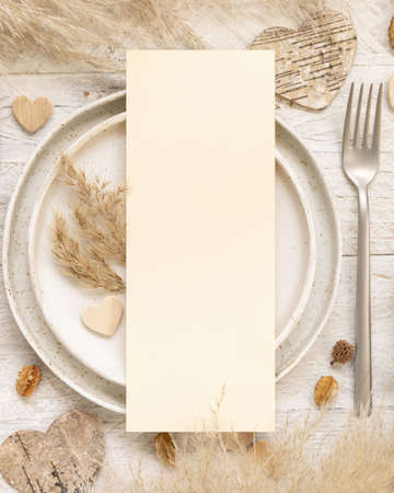 Wedding table place with blank menu card  near dried plants, palm leaves, pampas grass and hearts top view. Boho scene with vertical paper card flat lay. Valentines mockupの写真素材