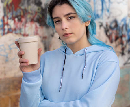 Teenage girl in light blue oversize hoodie with coffee to go and looking aside. Blue haired teen girl staying outdoors near graffiti wall. Clothing mockup.  Hipster and adolescence conceptの写真素材