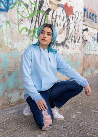 Blue haired Teenage girl in light blue oversize hoodie and looking aside. Blue haired teen girl sitting on her haunches near graffiti wall. Clothing mockupの写真素材