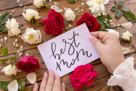Hands with handwritten card BEST MOM surrounded by red and cream roses close up on a wooden table. Femminine romantic declaration of love near flowers, Valentines day conceptの写真素材
