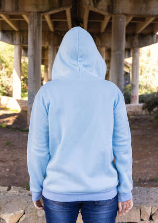 Blue haired Teenage girl in light blue oversize hoodie stand with her back to camera. Blue haired teen girl stays outdoors against bridge pillars. Clothing mockupの写真素材