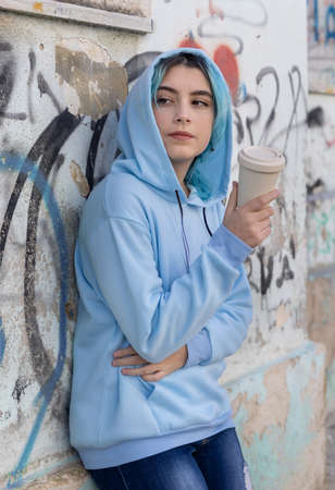 Teenage girl in light blue oversize hoodie with coffee to go and looking aside. Blue haired teen girl staying outdoors near graffiti wall. Clothing mockup.  Hipster and adolescence conceptの写真素材