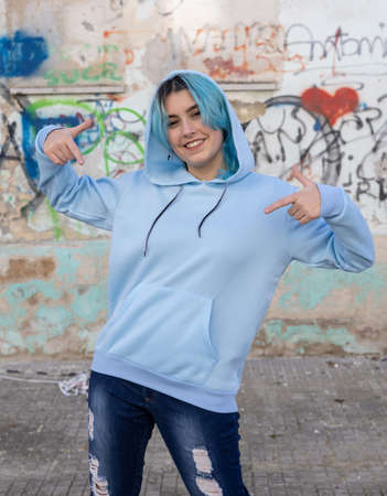 Smiling blue haired Teenager pointing in center of blue hoodie. Hooded teen girl stays outdoors against graffiti wall. Clothing mockupの写真素材