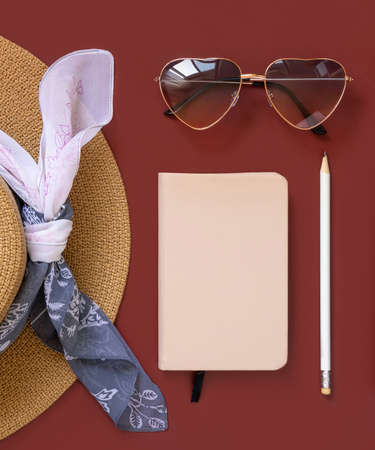 Straw hat, notebook, pensil and heart sunglasses top view on red background. Female summer clothes and accessories flat lay. Hardcover textbook mockup. Travel conceptの写真素材