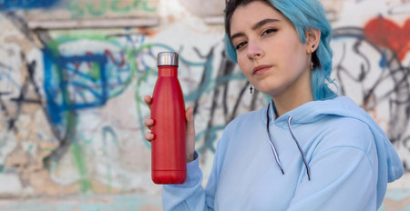 Teenager in blue hoodie with red water bottle looking stright. Blue haired teen girl staying...