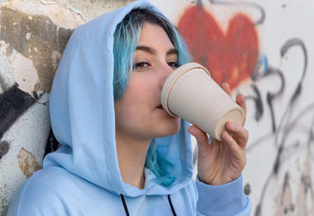Teenage girl in light blue oversize hoodie drinking coffee to go. Blue haired teen girl outdoors near graffiti wall. Coffee cup mockup. Hipster and adolescence concept. Head shotの写真素材