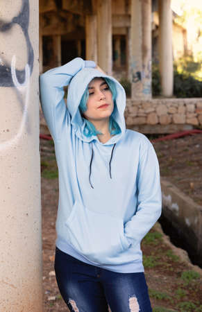 Smiling Teenager wearing light blue oversize hoodie and jeans stays under bridge . Blue haired teen girl stays outdoors against bridge pillars. Clothing mockupの写真素材