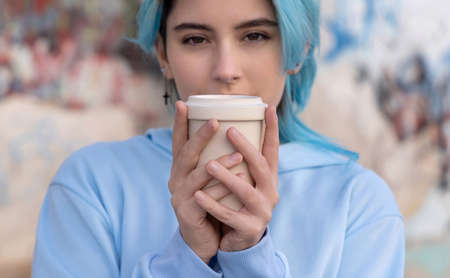 Teenage girl in light blue oversize hoodie with coffee to go and looking stright.Blue haired teen girl staying outdoors near graffiti wall, head shot.  Hipster and adolescence conceptの写真素材