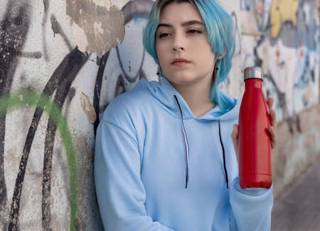 Teenager in blue hoodie with red water bottle looking aside. Blue haired teen girl staying outdoors near graffiti wall. Clothing and reusable bottle mockup.  Hipster and adolescence conceptの写真素材