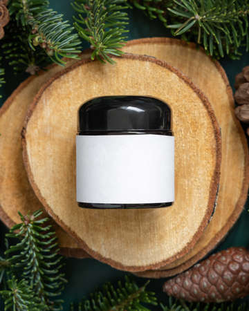 Glass jar between fir branches and pine cones on wooden piece top view. Brand packaging mockup. Dark cream container for skincare product, cream or serum. Healthcare concept. Natural cosmetics.の写真素材