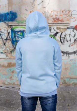 Young woman in blue hoodie stand with her back to camera.  Adolescent girl stays outdoors in front of graffiti wall. Clothing mockupの写真素材