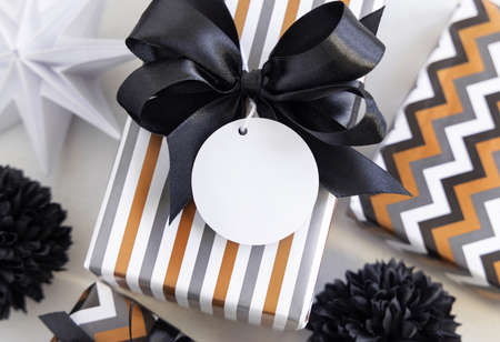Present with round blank gift tag and black bow close up. Gift boxes wrapped in Striped geometric paper near black and white decor. Christmas, New Year, Birthday, Anniversary label mockupの写真素材