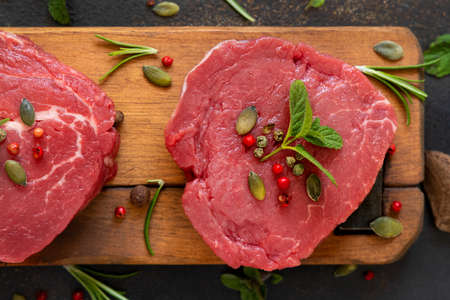 Raw beef fillet steaks on wooden board on dark table close up. Portioned filet mignons with spices and herbs. Concept of summer foodの写真素材
