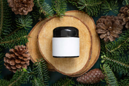 Glass jar between fir branches and pine cones on wooden piece top view. Brand packaging mockup. Dark cream container for skincare product, cream or serum. Healthcare concept. Natural cosmetics.の写真素材