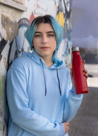 Teenager in blue hoodie with red water bottle looking stright. Blue haired teen girl staying outdoors near graffiti wall. Clothing and reusable bottle mockup.  Hipster and adolescence conceptの写真素材