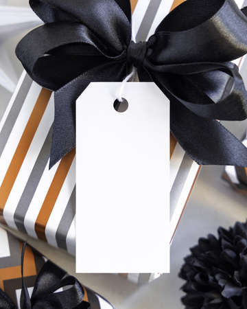 Present with vertical blank gift tag and black bow close up. Gift boxes wrapped in Striped geometric paper near black and white decor. Christmas, New Year, Birthday, Anniversary label mockupの写真素材