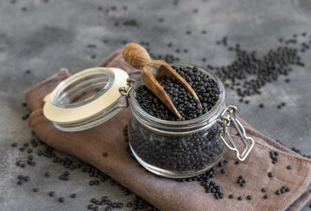 Glass jar of dry black lentils beans with a wooden scoop on grey table close up.   Healthy eating, protein source for vegan and vegetarian diet. Traditional Asian and North African cuisines ingredientの写真素材
