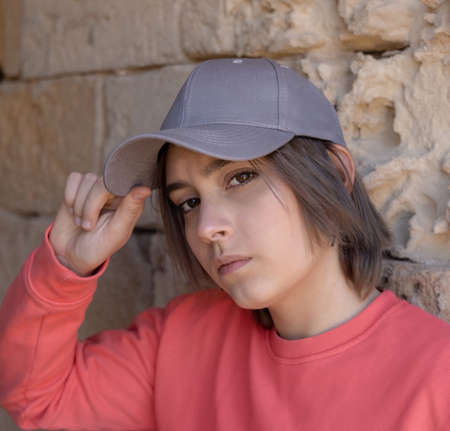 Teen girl in grey baseball cap and pink long sleeve shirt looking into a camera. Cap mockup. Serious Teenager girl near a stone wall outdoor touching the visor. Loneliness conceptの写真素材