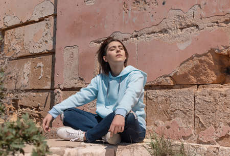 Teenager girl in light blue hoodie sitting on her haunches with eyes closed near a wall. Serious Teen girl spending time outdoor alone. Clothing mockup. Adolescence conceptの写真素材