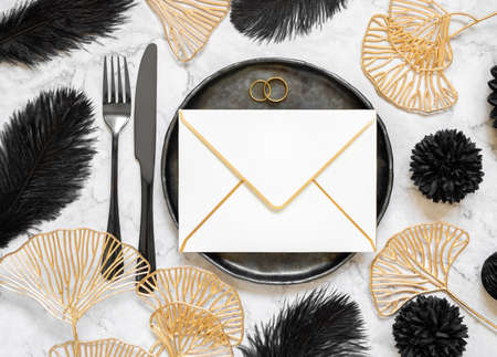 Black and golden wedding table setting with a blank envelope and rings top view, mockup. Elegant flat lay with horizontal greeting or Invitation paper envelope, wedding or holiday stationery templateの写真素材