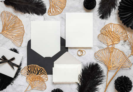 Blank cards and black envelope on marble table near black feathers and golden leaves top view. Wedding set mockup. Elegant femminine suite with paper cards, flat layの写真素材