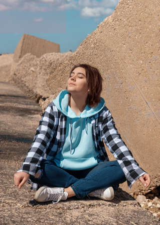 Happy Teenager girl in light blue hoodie and plaid shirt. Smiling Teen girl sitting outdoor cross-legged and enjoying the sunny day. Adolescence conceptの写真素材