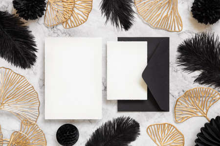 Blank cards and black envelope on marble table near black feathers and golden leaves top view. Wedding set mockup. Elegant femminine suite with paper cards, flat layの写真素材