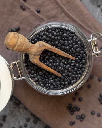 Glass jar of dry black lentils beans with a wooden scoop on grey table top view.   Healthy eating, protein source for vegan and vegetarian diet. Traditional Asian and North African cuisines ingredientの写真素材