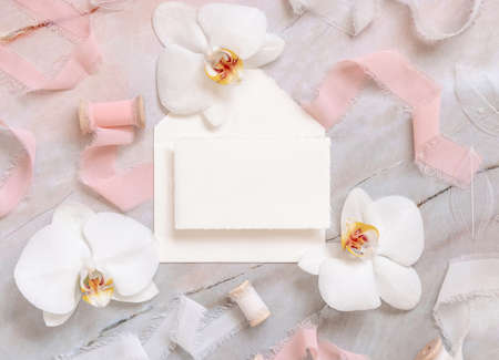 Wedding horizontal card and envelope near white orchid flowers and silk ribbons on marble top view, mockup. Romantic scene flat lay with handmade paper cardの写真素材
