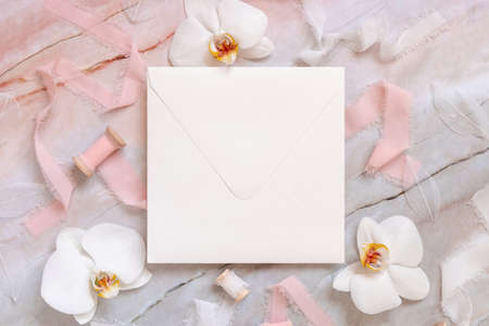 Wedding square envelope near white orchid flowers and silk ribbons on marble top view, mockup. Romantic scene flat lay. Valentines or Spring romantic conceptの写真素材