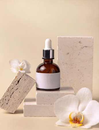Brown glass dropper bottle on stone near white orchid flowers on light yellow, close up, mockup. Skincare handmade beauty product, serum or lotion. Exotic natural cosmeticsの写真素材