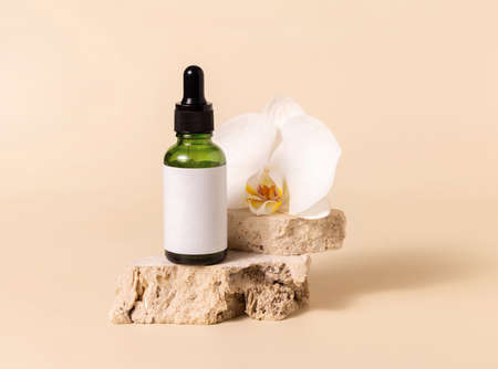 Green glass dropper bottle on stone near white orchid flower on light yellow, close up, mockup. Skincare handmade beauty product, serum or lotion. Exotic natural cosmeticsの写真素材