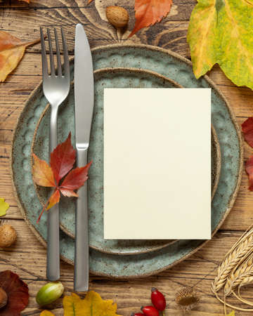 Autumn rustic table setting with blank card between colorful leaves and berries, top view. Vertical card mockup on wooden table, flat lay, copy space. Thanksgiving, halloween or harvest conceptの写真素材